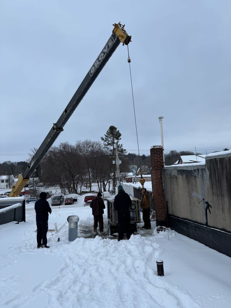 Technicians on snowy day installing large commercial HVAC unit with crane support