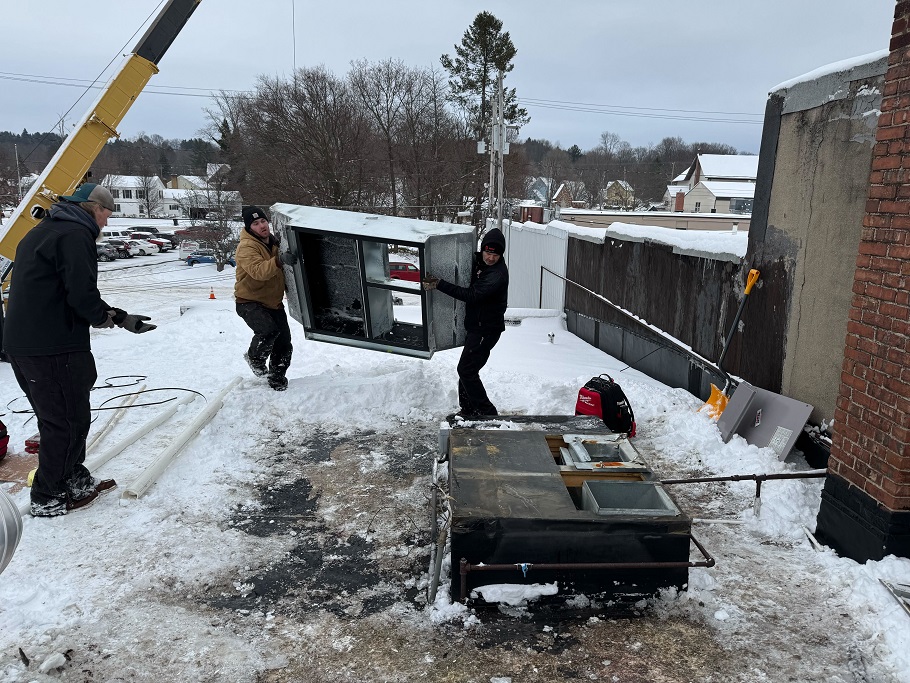 Technicians on snowy day installing large commercial HVAC unit with crane support