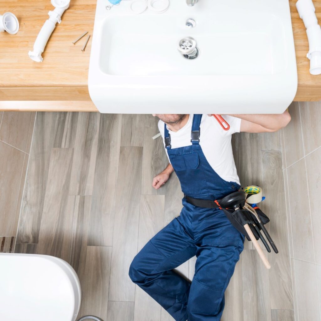 Plumber in blue overalls working under bathroom sink with tools and parts nearby