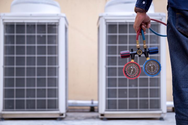 Hand holding HVAC manifold gauge set in front of two outdoor AC units
